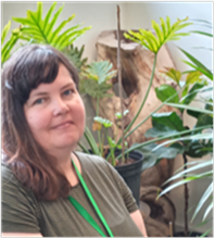 Photograph of Jenn Estepp, who is a white woman who has medium length brown hair and is wearing a brown shirt. There are houseplants in the background.